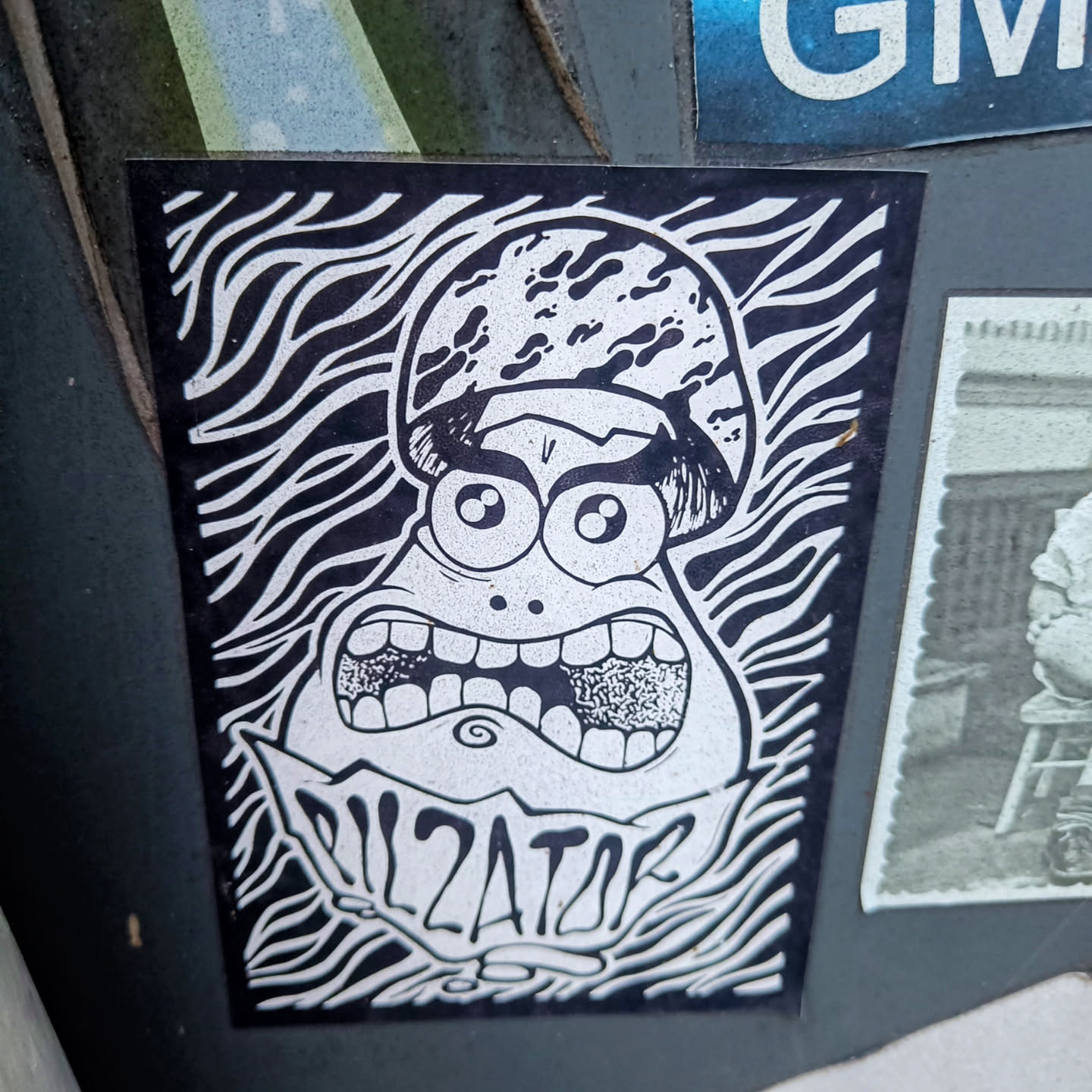 Black and white sticker of a cartoon character with big eyes and a wide open mouth. The character is wearing a helmet and has the word "DILATOR" written below it. The background is made up of wavy lines.