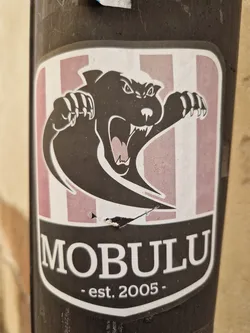 A sticker featuring a stylized black panther or similar creature with sharp claws, set against a background of pink and white vertical stripes.  The name "MOBULU" is prominently displayed below the image, along with the establishment date "est. 2005". The sticker appears to be affixed to a dark-colored pole.