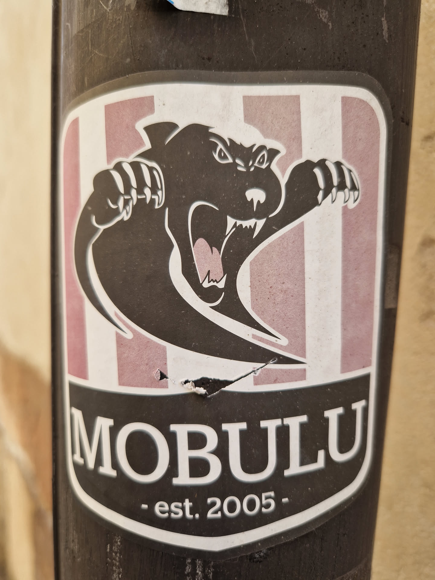 A sticker featuring a stylized black panther or similar creature with sharp claws, set against a background of pink and white vertical stripes.  The name "MOBULU" is prominently displayed below the image, along with the establishment date "est. 2005". The sticker appears to be affixed to a dark-colored pole.