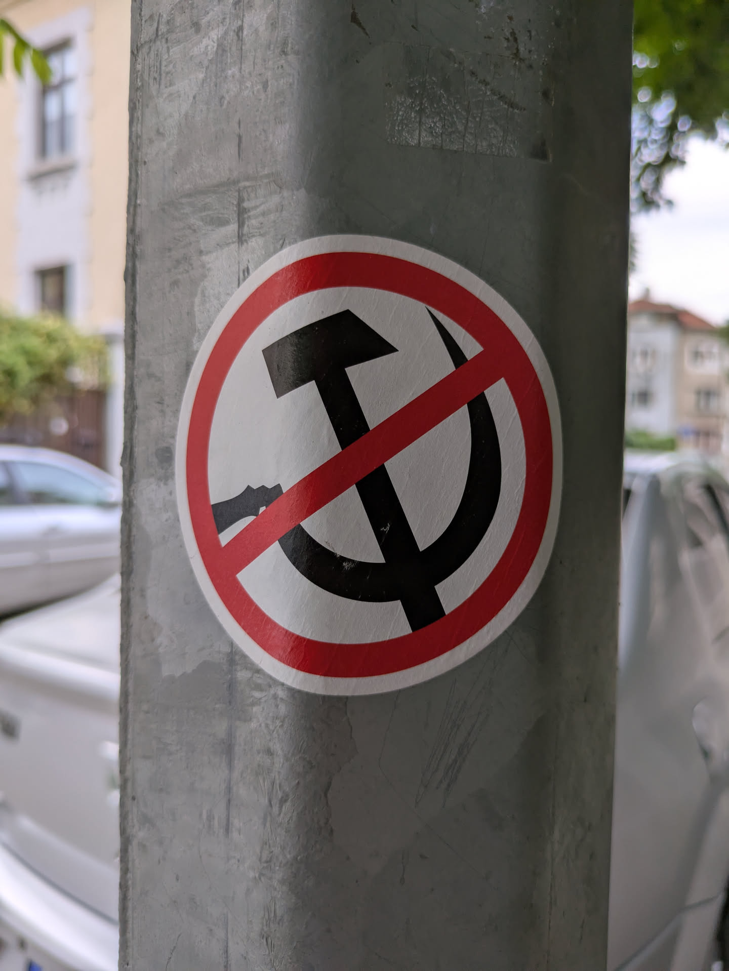 A round sticker adhered to a gray metal post features a black hammer and sickle symbol crossed out with a red circle and diagonal line. The red circle has a white interior.