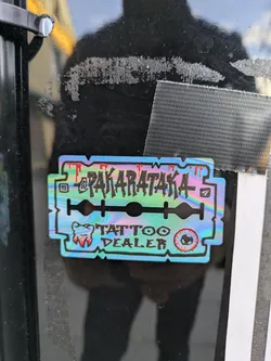 Holographic sticker in the shape of a razor blade featuring the text "Pakarataka" and "Tattoo Dealer". It also includes a small illustration of a tooth.