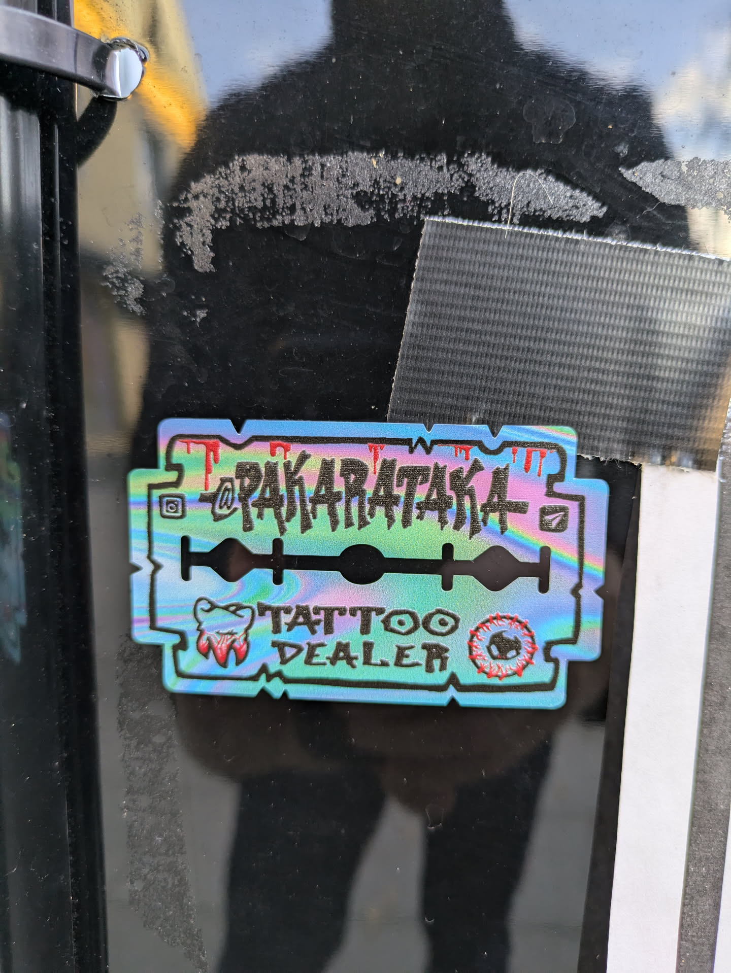 Holographic sticker in the shape of a razor blade featuring the text "Pakarataka" and "Tattoo Dealer". It also includes a small illustration of a tooth.