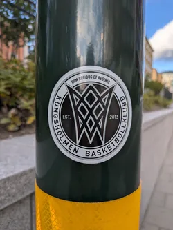 The sticker is circular with a white border. The central design features a stylized, abstract representation of a basketball hoop and net, rendered in black and white with sharp geometric lines. Inside the central design, the text 'EST.' is visible on the left, and '2013' is visible on the right. The outer ring of the sticker contains text in black capital letters on a white background, with a thin black outline. The text reads 'KUNGSHOLMEN BASKETBOLLKLUBB' along the bottom arc and 'CUM REGIBUS ET REGINIS' along the top arc.