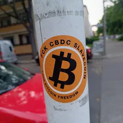 A round sticker featuring a Bitcoin symbol in the center, surrounded by the text: "FCK CBDC SLAVECOIN LOVE BITCOIN FREEDOM". The sticker is affixed to a white pole outdoors.