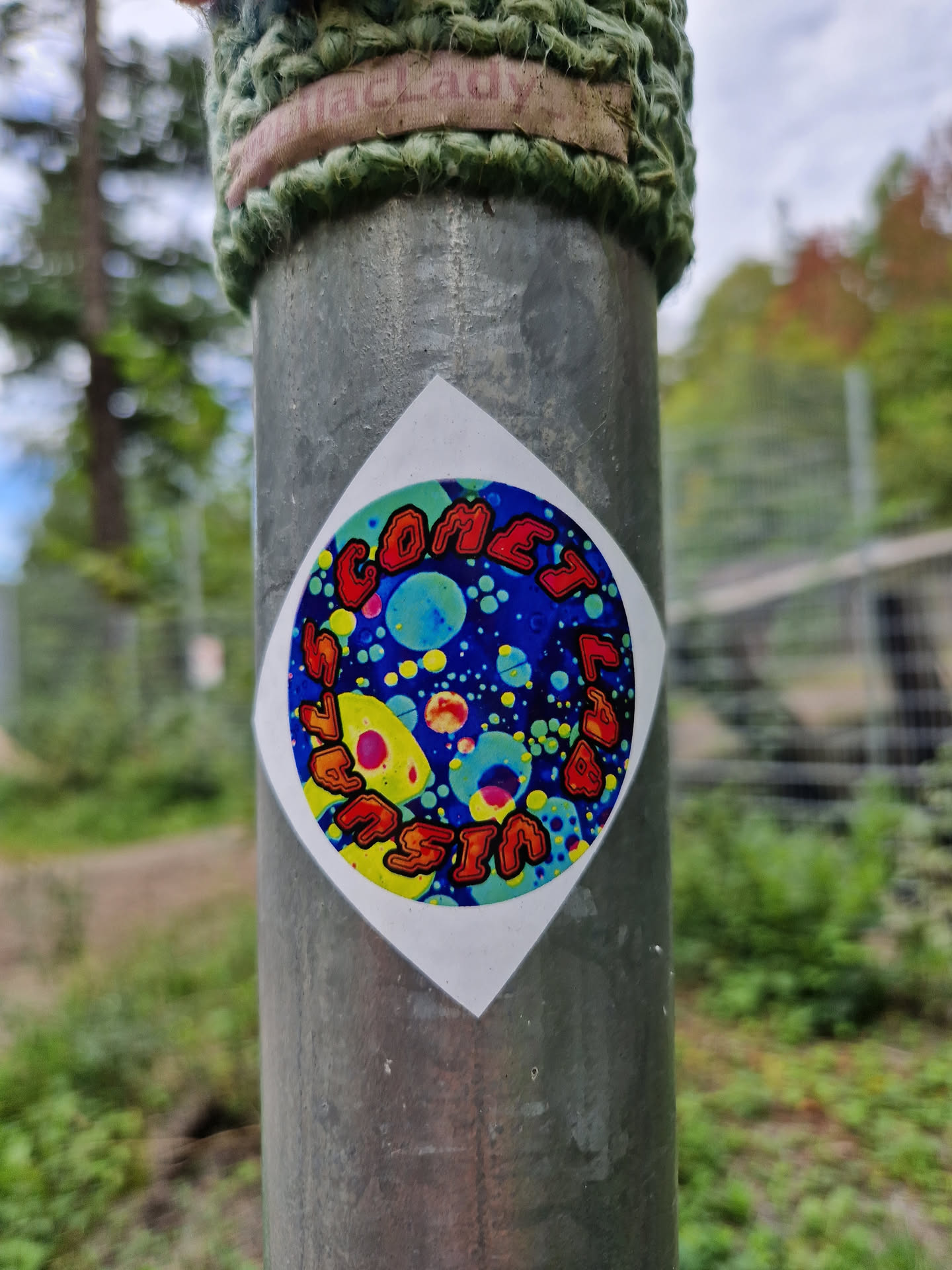 A colorful sticker featuring the words "Comet Season" in a stylized font, set against a background of swirling colors and bubbles. The sticker has a diamond shape with rounded corners and is affixed to a metal pole.