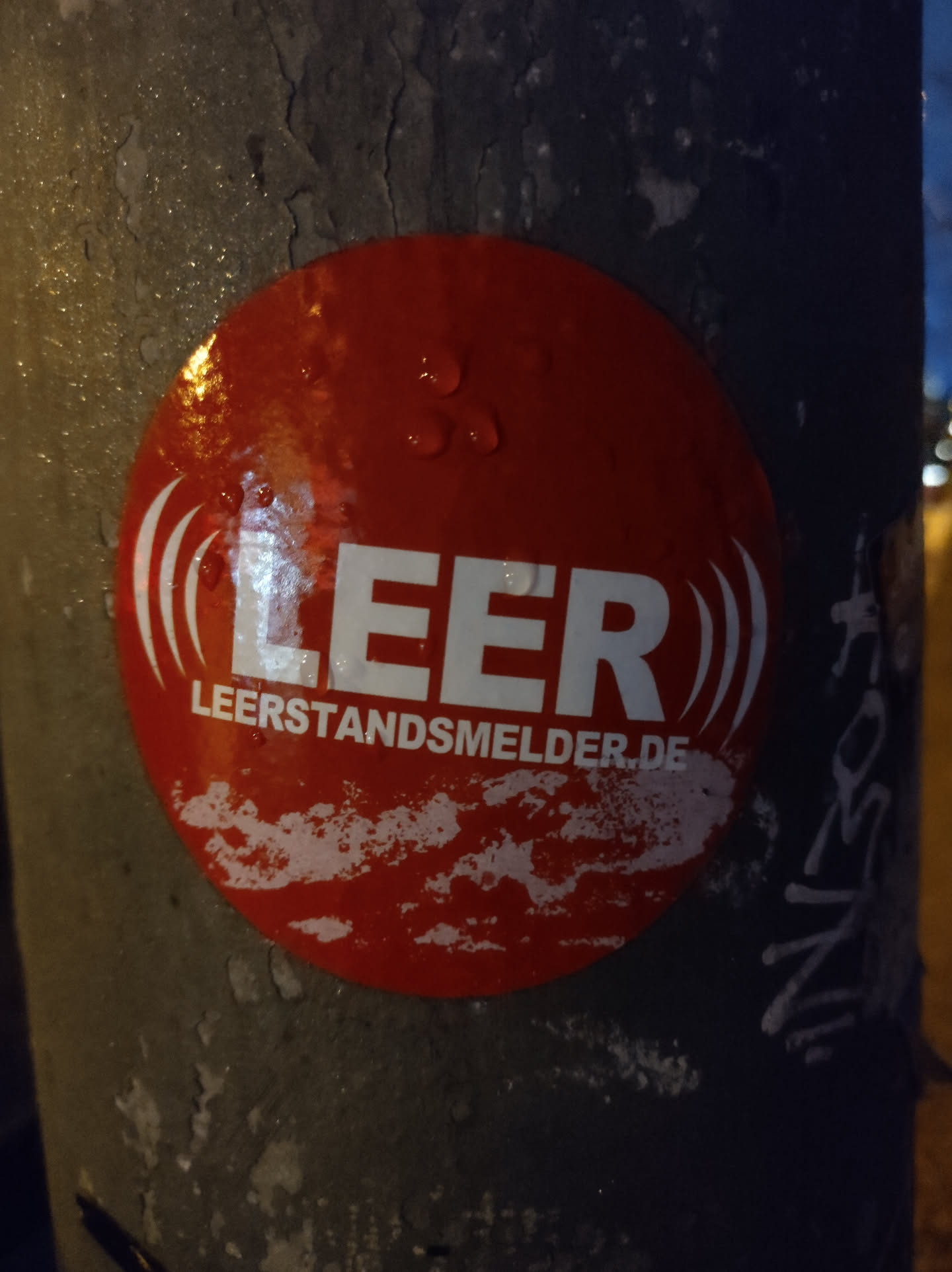 A red circular sticker is affixed to a gray pole. The sticker displays the word "LEER" prominently in white uppercase letters, with stylized sound wave symbols on either side.  Below "LEER" is the smaller text "leerstandsmelder.de". The sticker appears slightly damp, showing water droplets on its surface.
