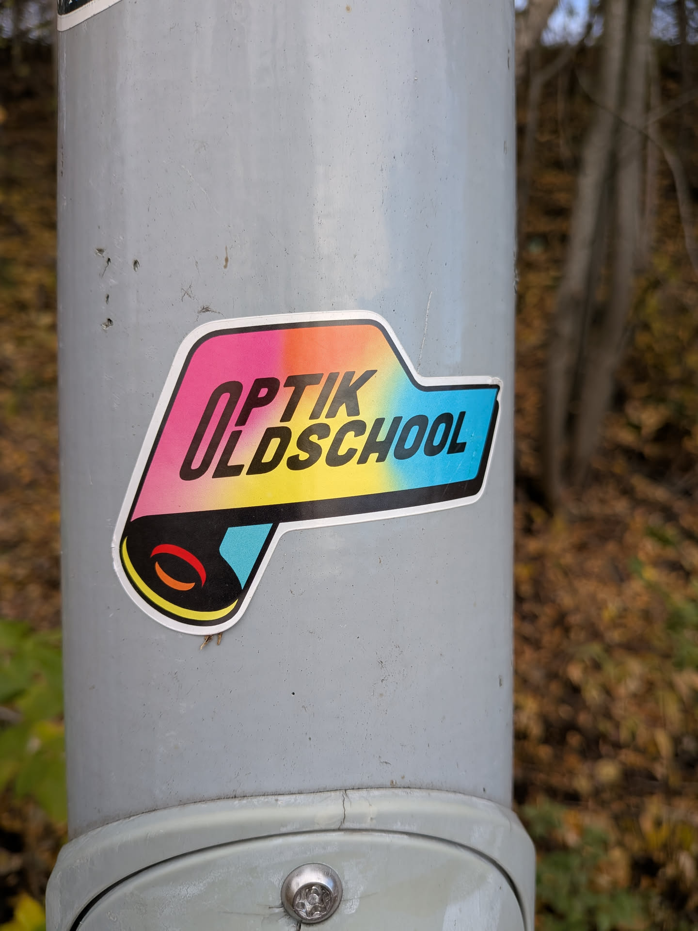 The sticker is centrally located and features a bold, stylized design. The main text reads 'OPTIK OLDSCHOOL' in black, sans-serif capital letters. The background of the text is a vibrant gradient transitioning from pink to orange, yellow, and then a bright cyan blue. The sticker has a white border and a black outline. Below the text, there's a graphic element resembling a rolled-up scroll or a film reel, with red and orange curved lines within a black circle, set against a cyan background. The overall shape of the sticker is somewhat irregular, with a pointed extension on the right side.