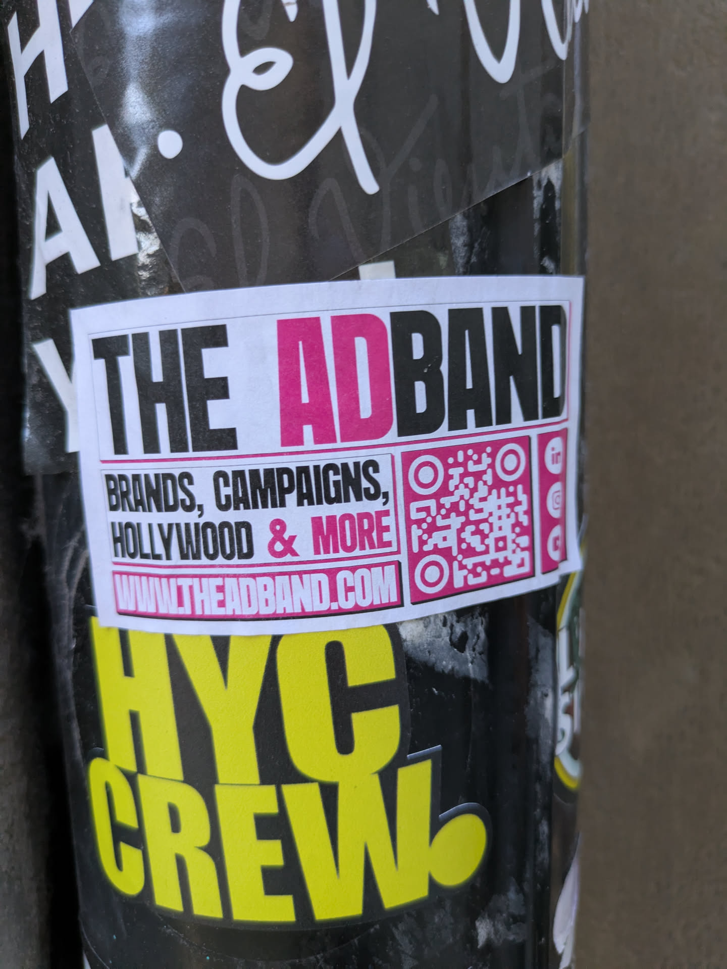 The sticker is rectangular with a white background and a pink border. At the top, bold black sans-serif letters spell out "THE AD" followed by "BAND" in bright pink. Below this, in smaller black text, are the words "BRANDS, CAMPAIGNS, HOLLYWOOD & MORE". Beneath that, centered and slightly larger, is a URL: "WWW.THEADBAND.COM". To the right of the text is a pink and white QR code. To the right of the QR code, there are three small circular icons that appear to be social media logos (LinkedIn, Instagram, and another unidentifiable one).