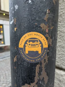 A round sticker with a dark-grey background and yellow accents featuring a jeep graphic and text. The sticker is affixed to a dark-grey, weathered pole.