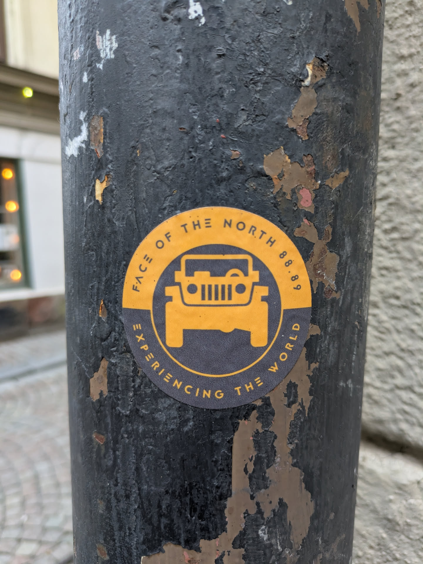 A round sticker with a dark-grey background and yellow accents featuring a jeep graphic and text. The sticker is affixed to a dark-grey, weathered pole.
