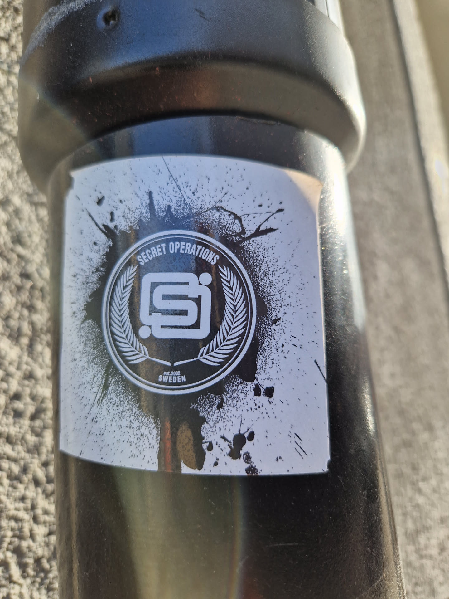 Black and white sticker with a logo and the words "Secret Operations" and "Sweden". The logo is a stylized "SI" inside a circle with laurel leaves, with a grunge effect around it.