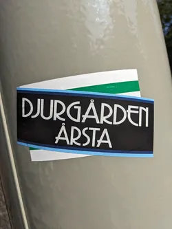 A sticker featuring the text "Djurgården Årsta" in a stylized art deco font.  The sticker is black with light blue and green accents.