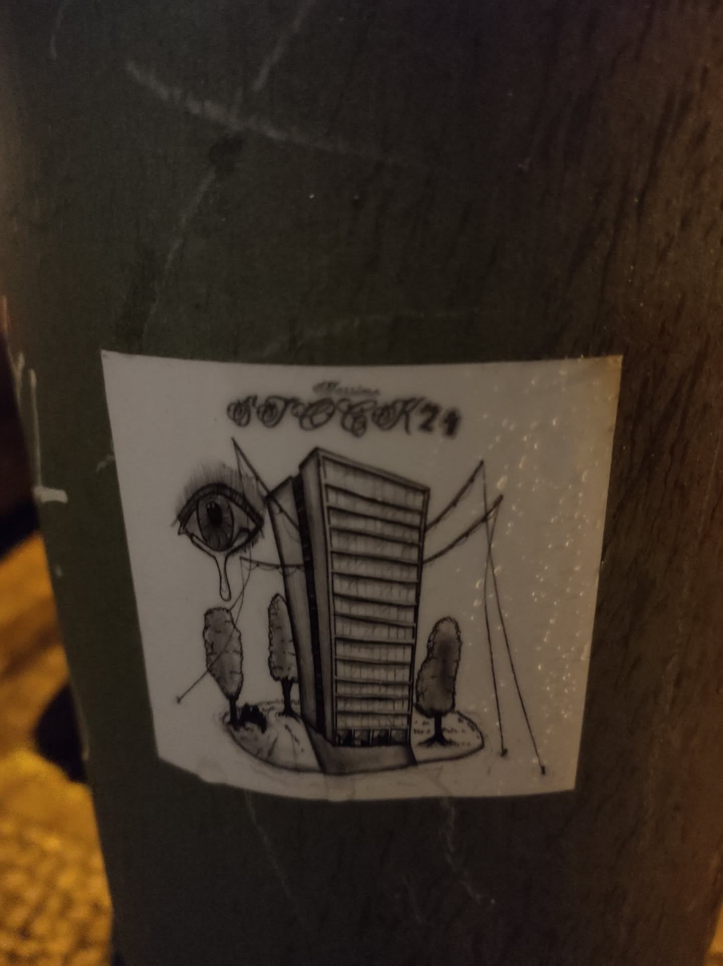 Black and white drawing of a tall building with small trees around it, and a crying eye watching over it. The style is surreal and somewhat melancholic. It's stuck on a pole outside.