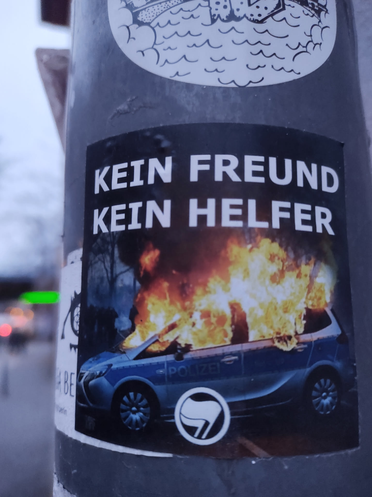 A sticker depicting a burning police car with the text "Kein Freund Kein Helfer" (No friend, no helper) and an anarchist symbol. 