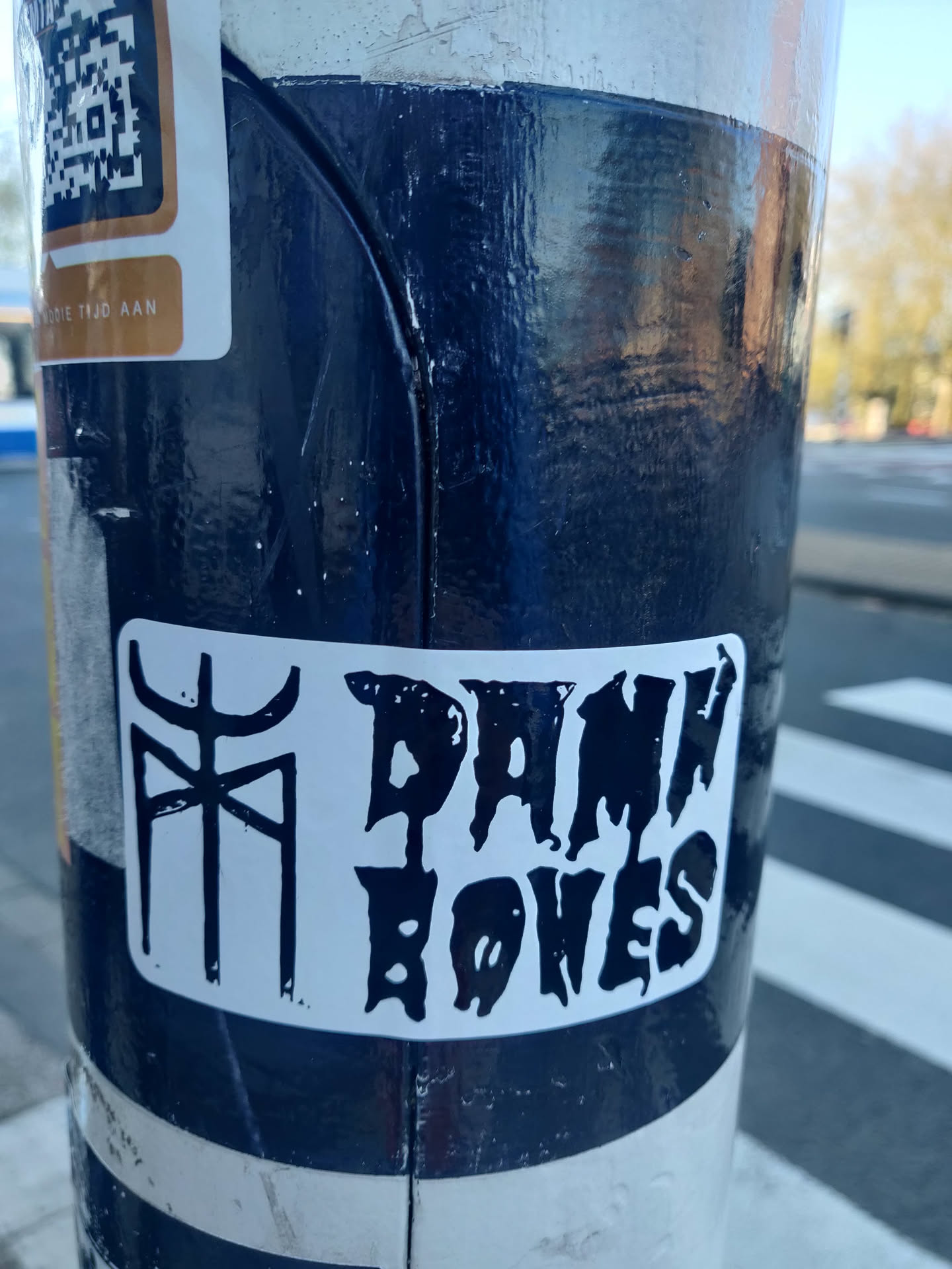 A black and white sticker with the text "Damn Bones" in a stylized font. There is also a smaller symbol to the left of the text. The sticker is affixed to a dark blue pole.