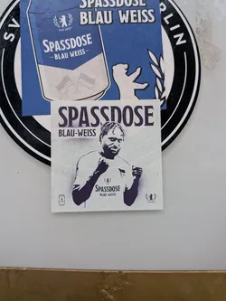 A square paper sticker rendered in dark navy ink on an off-white textured background. At the top, the bold header reads 'SPASSDOSE' above 'BLAU-WEISS'. The central image shows a stylized depiction of a smiling football player with dreadlocks, raising both fists in celebration. On his chest, a graphic replaces the shirt sponsor with 'SPASSDOSE -BLAU-WEISS-' next to the Hertha BSC flag emblem. A small line drawing of a beverage can appears at the bottom left, while a Berlin bear surrounded by a laurel wreath and the text 'SEIT 2023' is positioned at the bottom right.