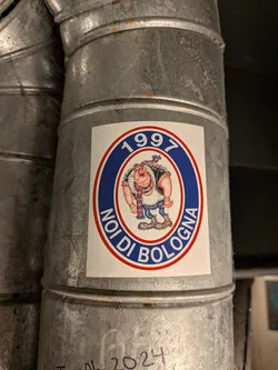Street sticker An oval-shaped sticker adhered to a galvanized metal ventilation duct. The sticker has a white rectangular background containing an oval crest outlined in red. The outer band of the oval is royal blue with white text reading '1997' at the top and 'NOI DI BOLOGNA' along the bottom. Inside the inner white oval is a cartoon caricature of a muscular, aggressive football hooligan with a prominent jaw, wearing a studded black vest, spiky boots, blue jeans, wristbands, and a red-and-blue striped scarf associated with Bologna FC colors.
