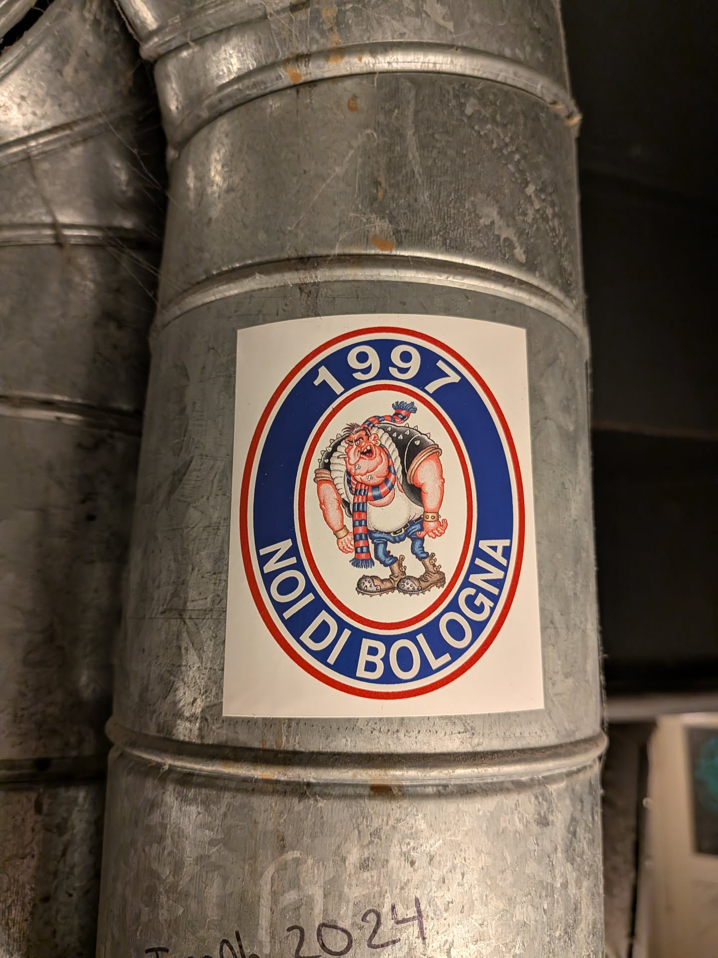 An oval-shaped sticker adhered to a galvanized metal ventilation duct. The sticker has a white rectangular background containing an oval crest outlined in red. The outer band of the oval is royal blue with white text reading '1997' at the top and 'NOI DI BOLOGNA' along the bottom. Inside the inner white oval is a cartoon caricature of a muscular, aggressive football hooligan with a prominent jaw, wearing a studded black vest, spiky boots, blue jeans, wristbands, and a red-and-blue striped scarf associated with Bologna FC colors.