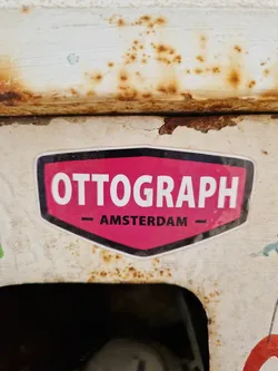 A pink and black sticker with the word OTTOGRAPH and AMSTERDAM written on it. The sticker is attached to a rusty surface.