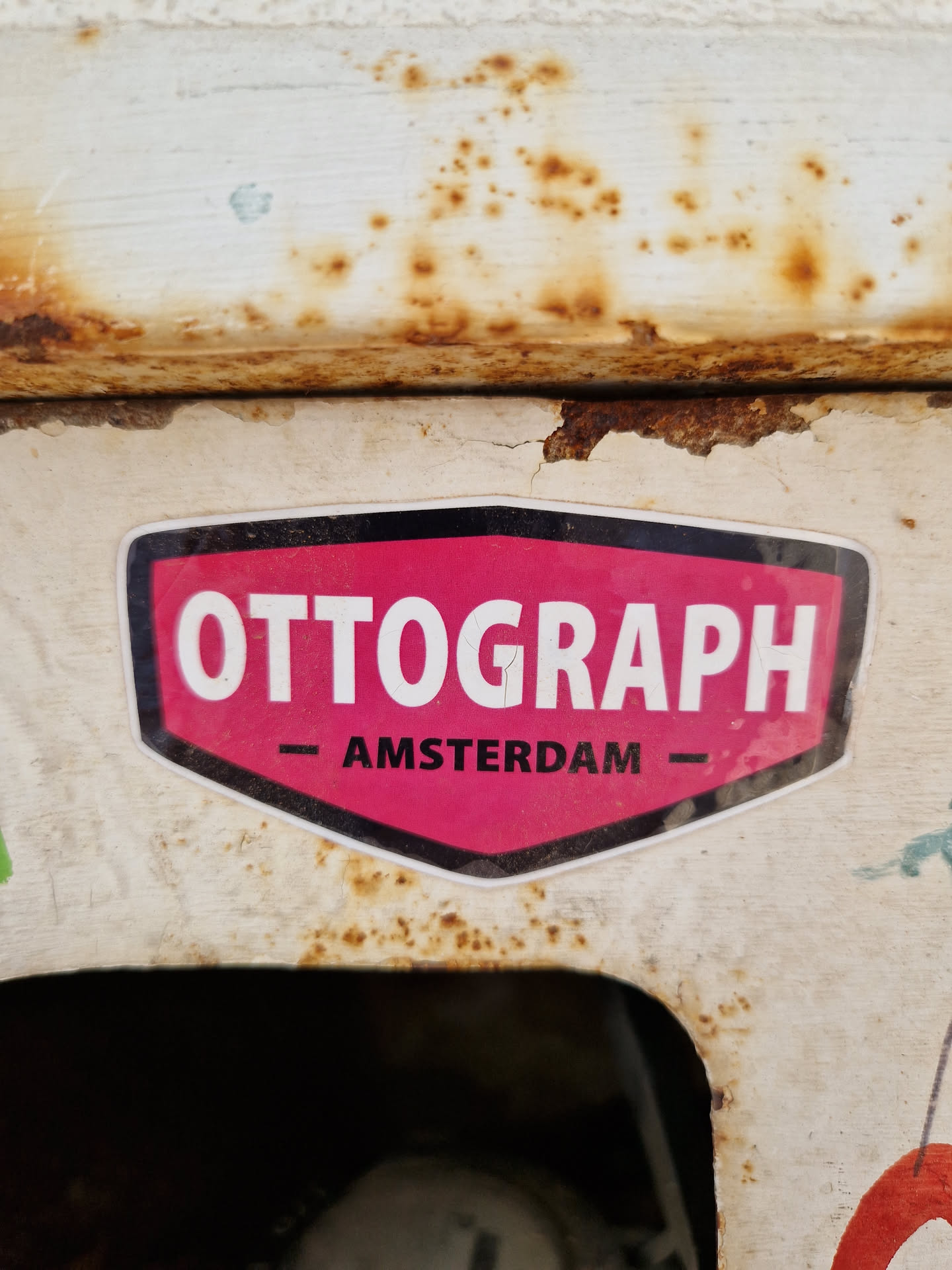 A pink and black sticker with the word OTTOGRAPH and AMSTERDAM written on it. The sticker is attached to a rusty surface.