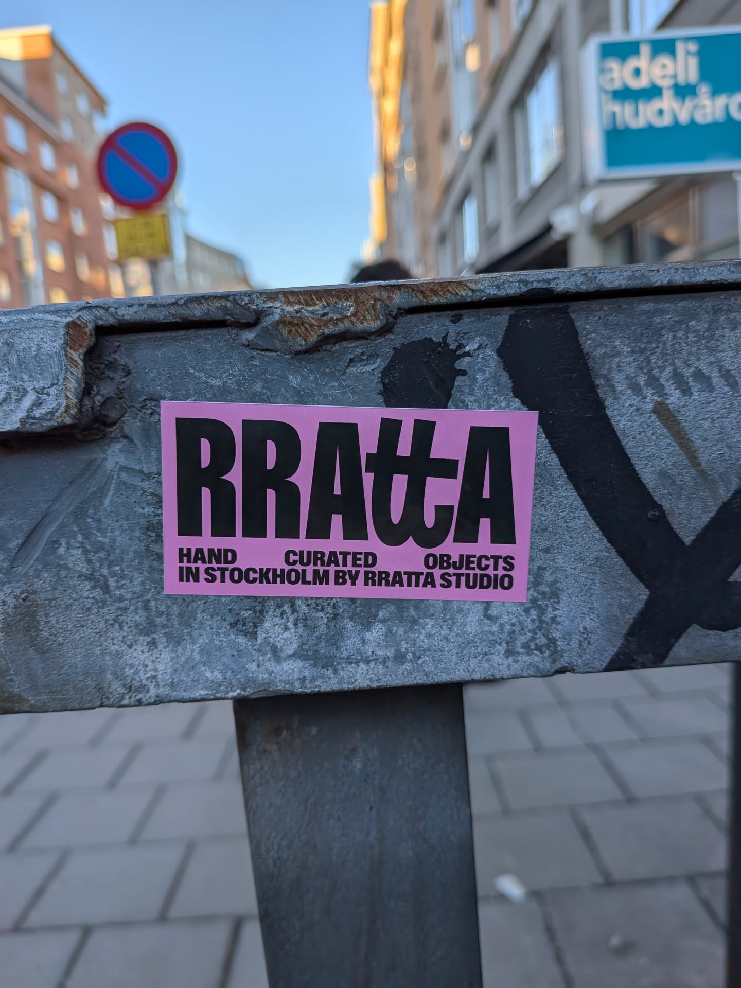 A rectangular sticker with a vibrant, bright pink background. The main text, 'RRATTA', is prominently displayed in large, bold, black sans-serif letters. The 'A' in 'RRATTA' is stylized, resembling a Greek letter pi (π) or a unique ligature of 'tt' and 'a'. Below this, in smaller black sans-serif text, are the words 'HAND CURATED OBJECTS'. Further below, in even smaller black sans-serif text, it reads 'IN STOCKHOLM BY RRATTA STUDIO'. The sticker is affixed to a weathered, rusty grey metal beam, with some black graffiti marks visible around it.