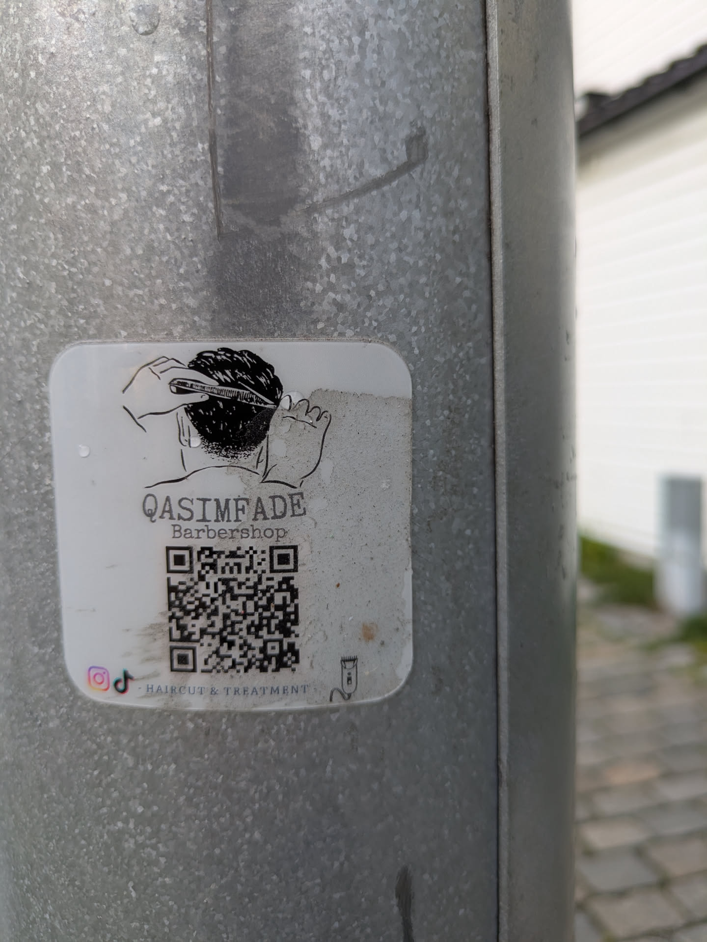 A white, rounded square sticker is affixed to a textured metal pole. The sticker prominently displays a black and white line drawing depicting the back of a head receiving a haircut, with hands holding a comb and clippers. Below the illustration, the text 'QASIMFADE' is visible, followed by 'Barbershop' and 'HAIRCUT & TREATMENT'. A large QR code is centered on the sticker. At the bottom left, small Instagram and TikTok logos are present. A tiny illustration of a hair clipper is on the bottom right.