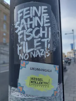 Two stickers are attached to a pole. The top sticker features the text "Feine Sahne Fischfilet, No Nazis" written in a stylized graffiti font. The bottom sticker is smaller and features the text "UKUMANGALA Kessel Kollektiv Stuttgart". The design of the second sticker includes decorative corner elements.