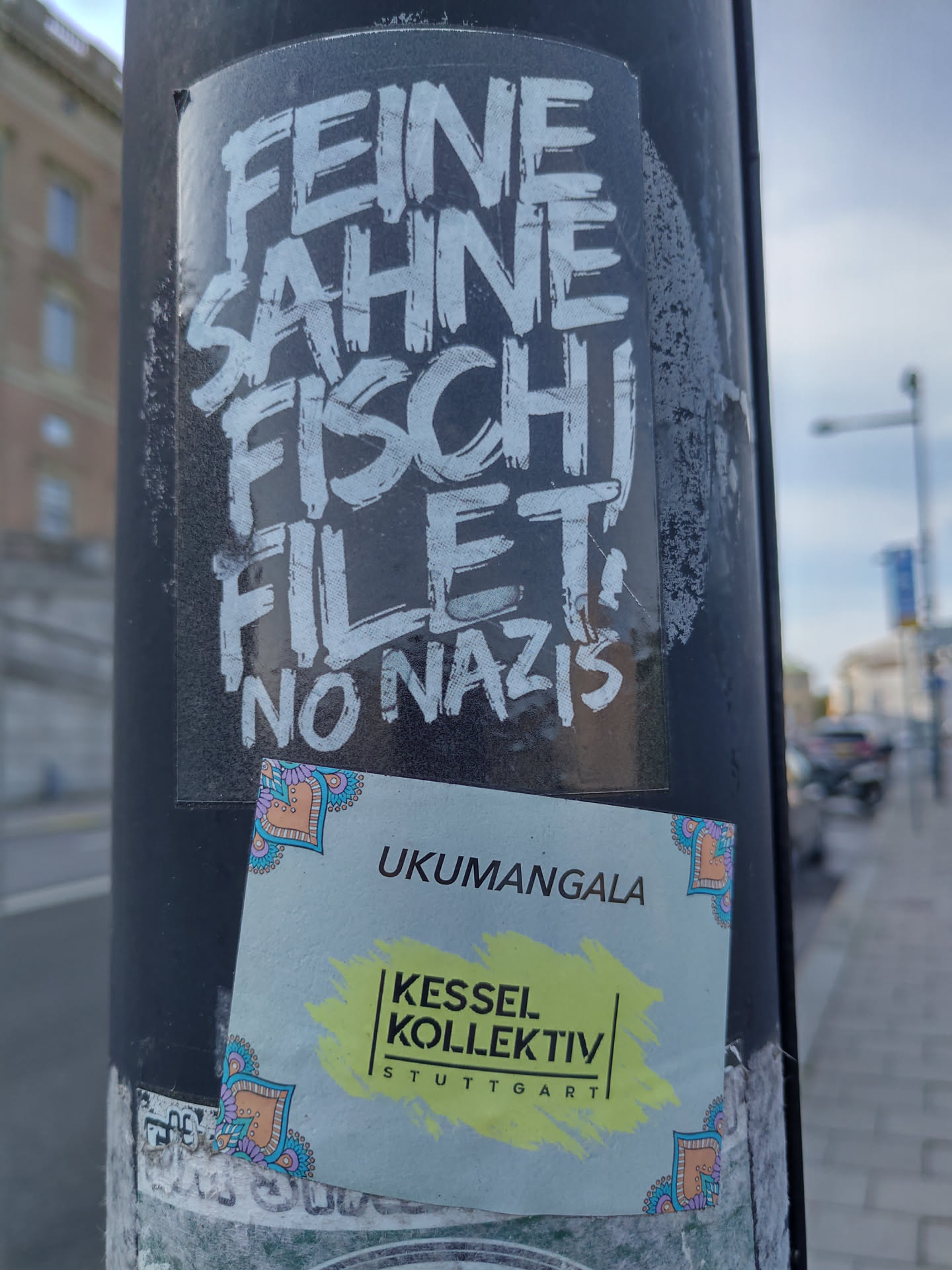 Two stickers are attached to a pole. The top sticker features the text "Feine Sahne Fischfilet, No Nazis" written in a stylized graffiti font. The bottom sticker is smaller and features the text "UKUMANGALA Kessel Kollektiv Stuttgart". The design of the second sticker includes decorative corner elements.