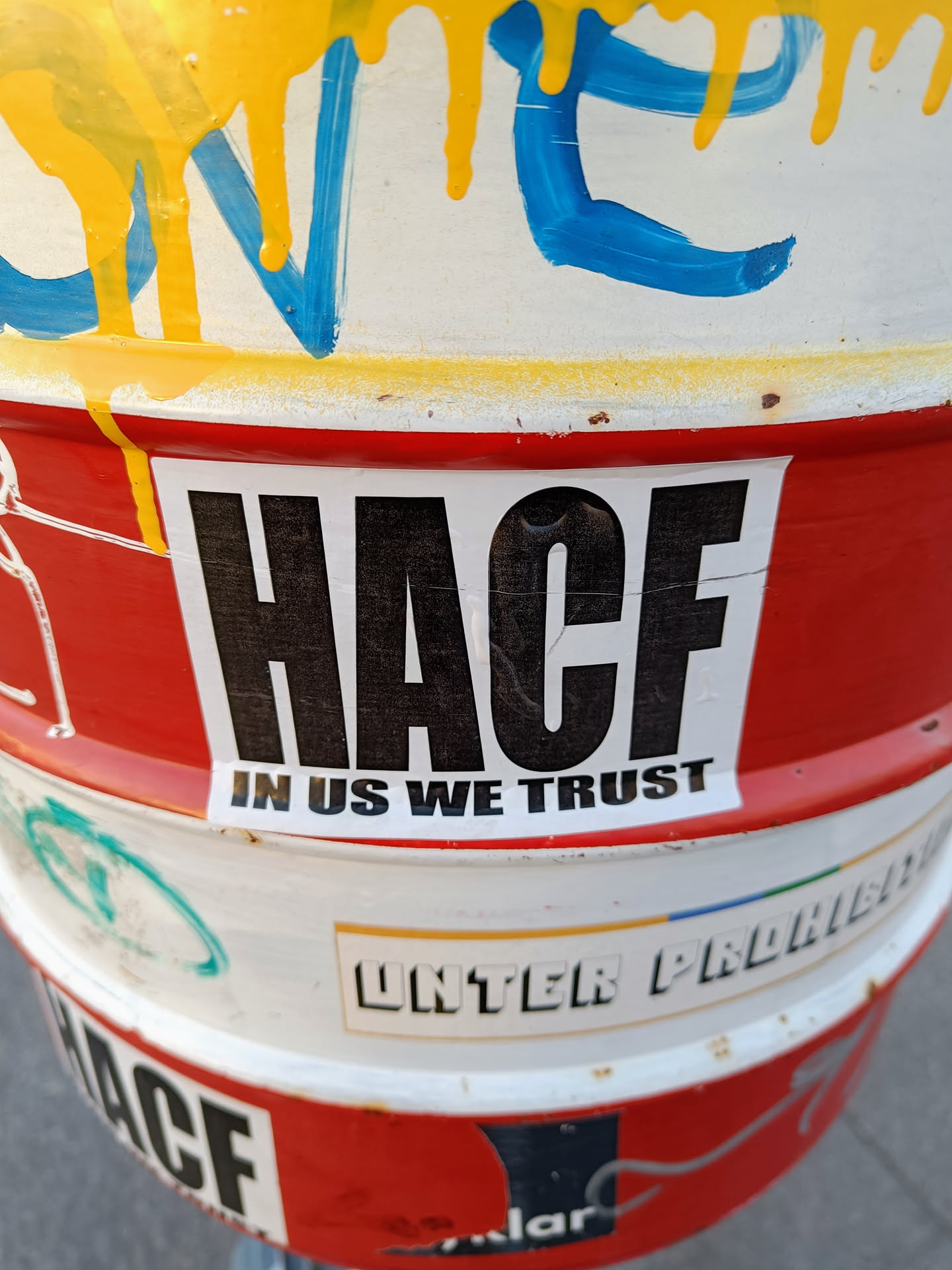 A red and white sticker with the text "HACF" in large, bold, black letters and the phrase "IN US WE TRUST" in smaller letters beneath. The sticker is affixed to a metal barrel that has other graffiti and stickers on it.