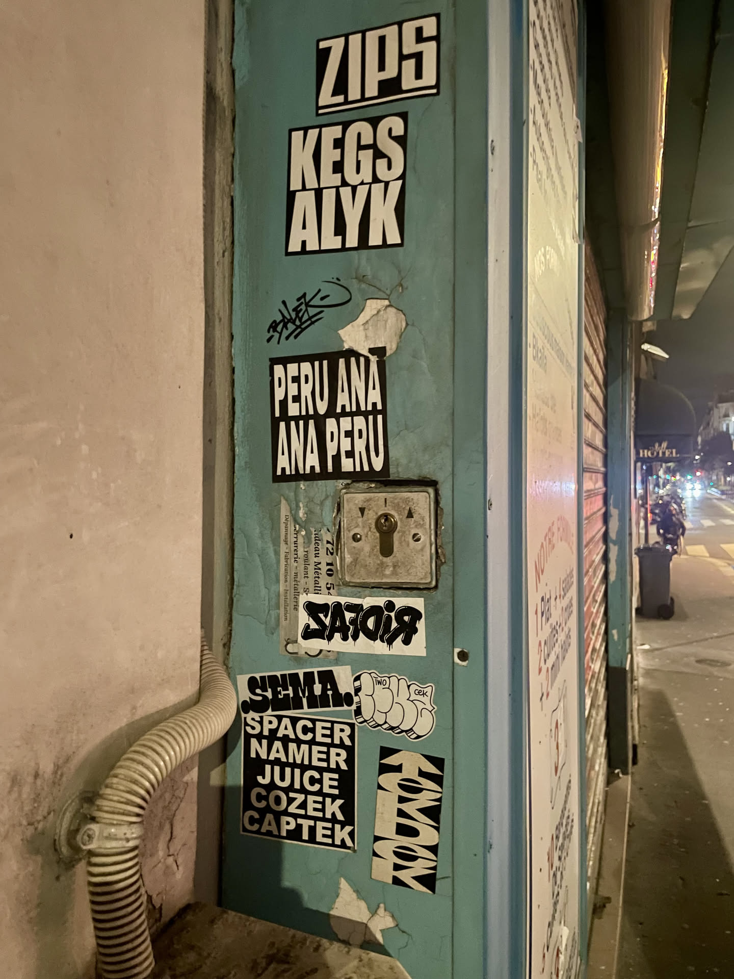 The sticker is a rectangular piece with a black background and bold, white, sans-serif capital letters. The text is arranged in two lines: 'PERU ANA' on the top and 'ANA PERU' on the bottom, creating a visually balanced or palindromic effect. The sticker shows signs of wear, with a noticeable tear and peeling at its top right corner, revealing the light blue-green surface it's adhered to. The overall appearance is stark and graphic, typical of street art tags.