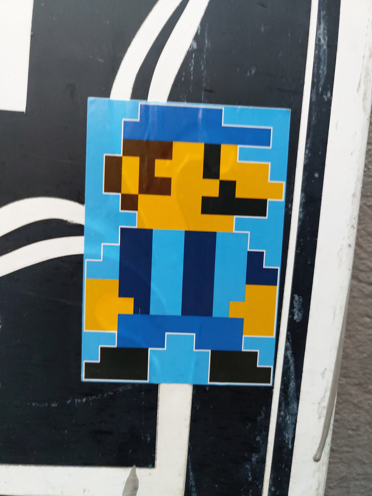 A sticker featuring a pixelated character resembling a video game character, predominantly in blue, yellow, and black. The character has a simple design with blocky features.