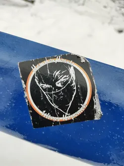 A worn sticker featuring a stylized illustration of a person's face partially obscured by a mask or cloth.  The design is encircled by a ring with text that reads "High rise Hoods" at the top and "witness keeper" at the bottom. The style is reminiscent of street art or graphic design.