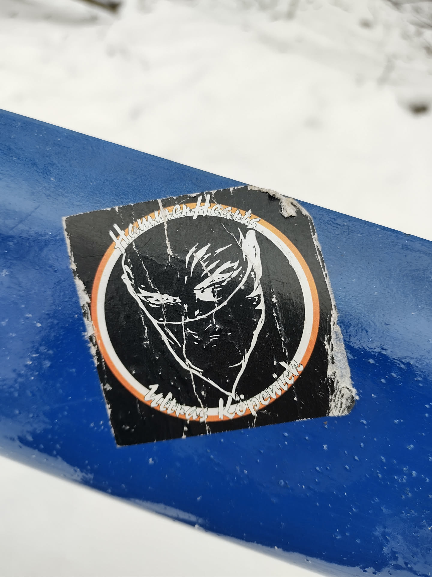 A worn sticker featuring a stylized illustration of a person's face partially obscured by a mask or cloth.  The design is encircled by a ring with text that reads "High rise Hoods" at the top and "witness keeper" at the bottom. The style is reminiscent of street art or graphic design.