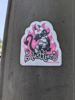 A white sticker with a cartoon illustration of a demon character designed by Belastiian. The demon is black and white, with a devil tail, small horns, wearing a hat and striped shirt. Pink flame-like shapes surround the character. The text "Belastiian" is written in a playful cursive font with their Instagram handle, @BELASTIIAN, below.