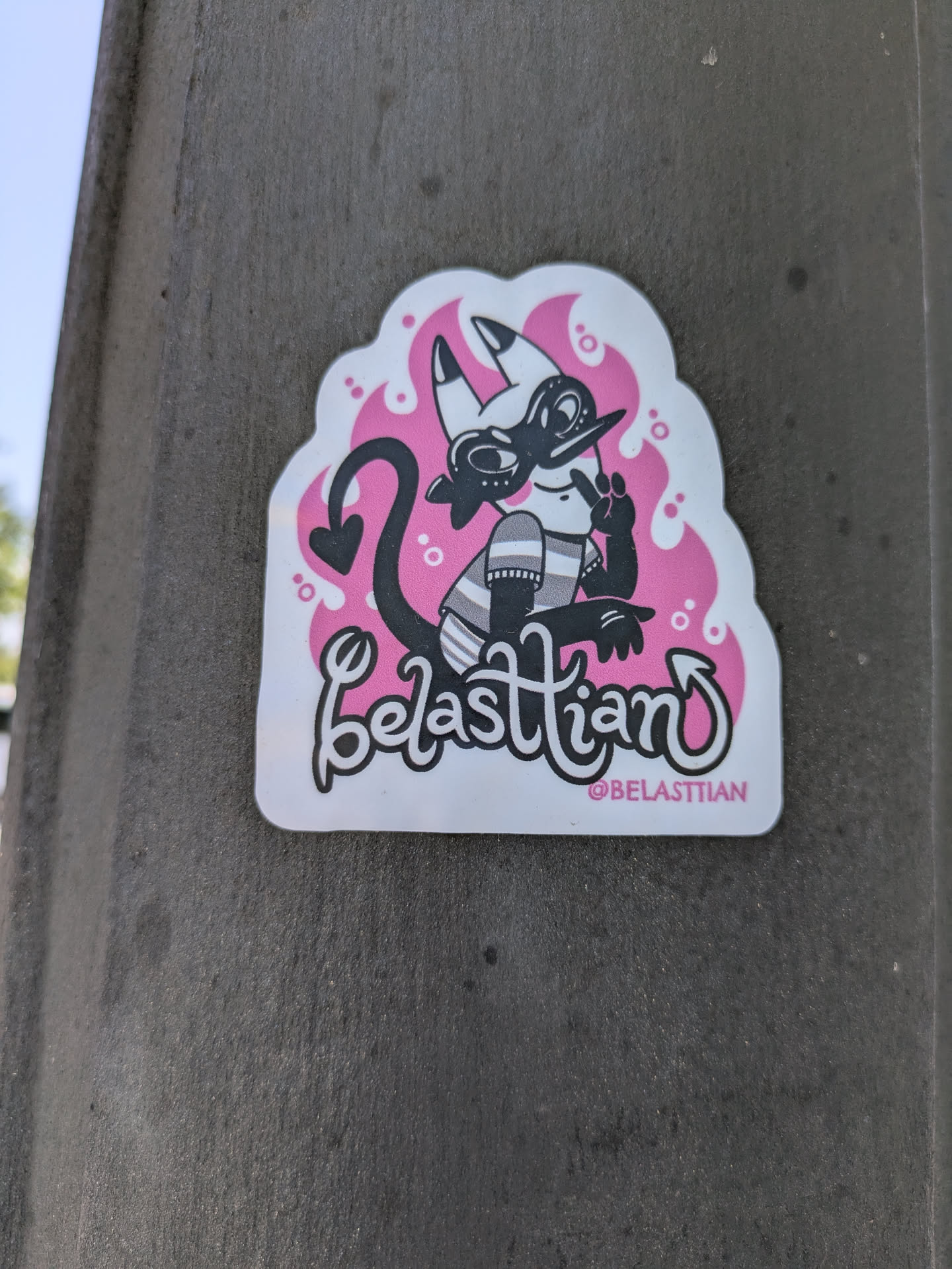 A white sticker with a cartoon illustration of a demon character designed by Belastiian. The demon is black and white, with a devil tail, small horns, wearing a hat and striped shirt. Pink flame-like shapes surround the character. The text "Belastiian" is written in a playful cursive font with their Instagram handle, @BELASTIIAN, below.