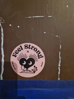 Pink round sticker with black text and illustration of a sad heart. Text on the sticker says "Feel strong" with "Odd Stories" and "Atelier" written smaller on the sides. 