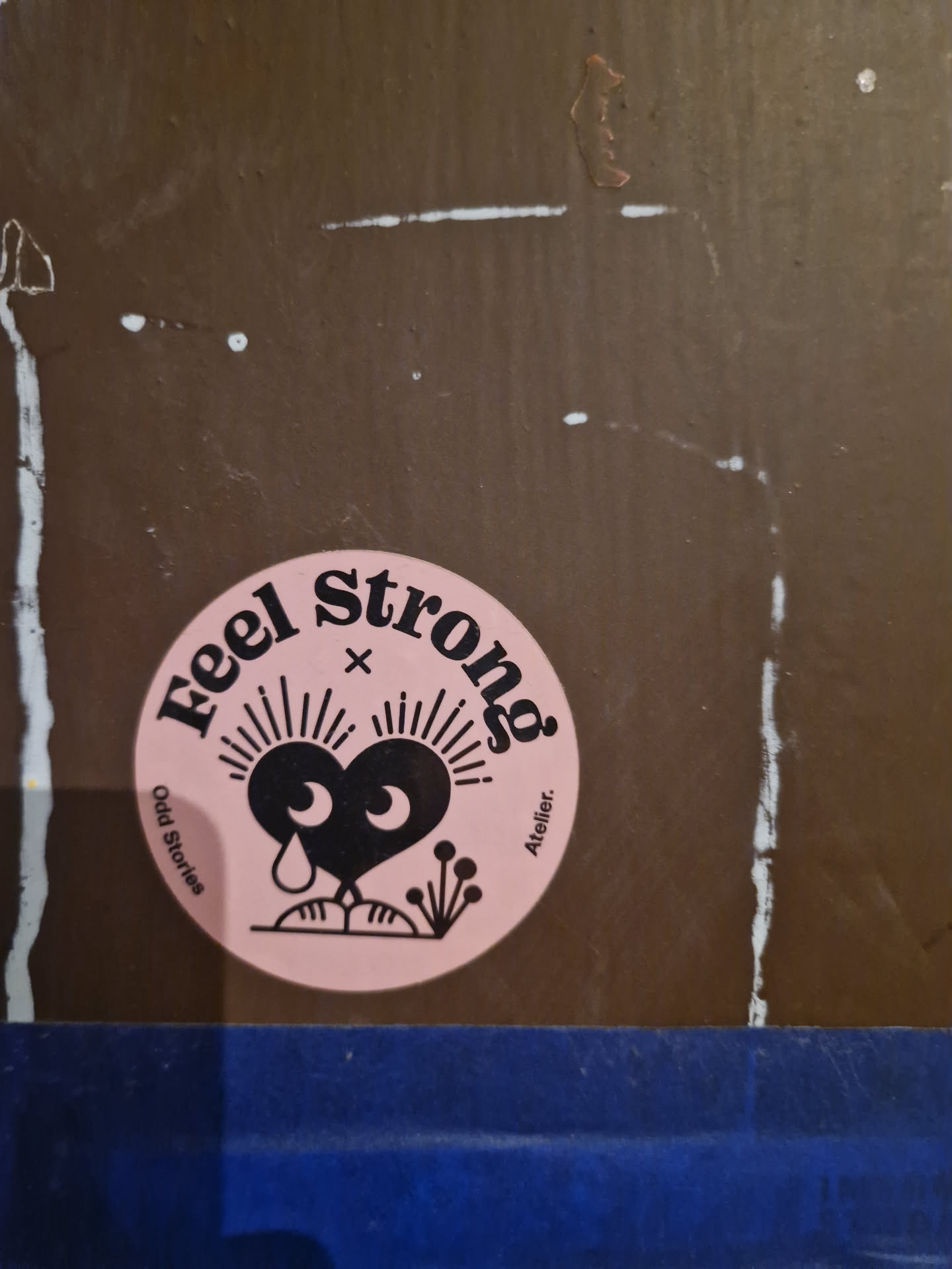 Pink round sticker with black text and illustration of a sad heart. Text on the sticker says "Feel strong" with "Odd Stories" and "Atelier" written smaller on the sides. 