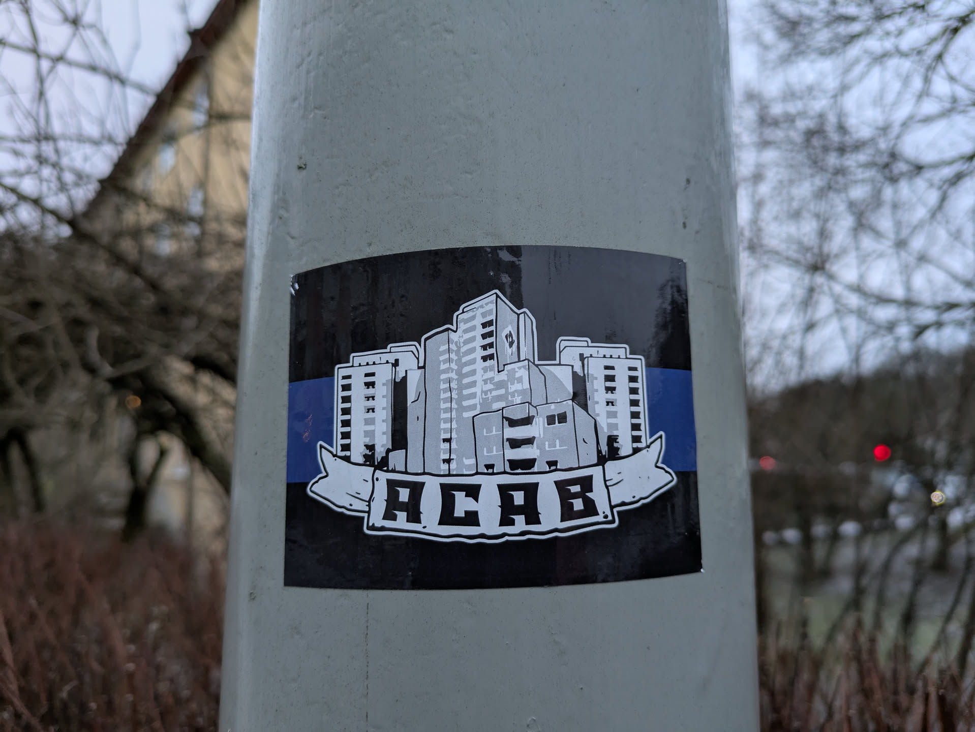 A sticker featuring a black and white illustration of apartment buildings, with a blue stripe at the top and bottom, and the acronym "ACAB" in a banner at the bottom. The sticker is affixed to a gray pole outdoors.