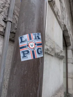 A square sticker placed on a brown painted metal pipe. It features the acronym 'LKPG' in stylized 3D serif block letters arranged across the corners on a light blue-grey field. Divided by a cross design composed of dark blue stripes bordered by white and red lines, the center bears the circular crest of Linköpings Hockey Club, displaying a roaring white lion head against a red center, encircled by a dark blue band reading 'LINKÖPINGS HOCKEY CLUB'.