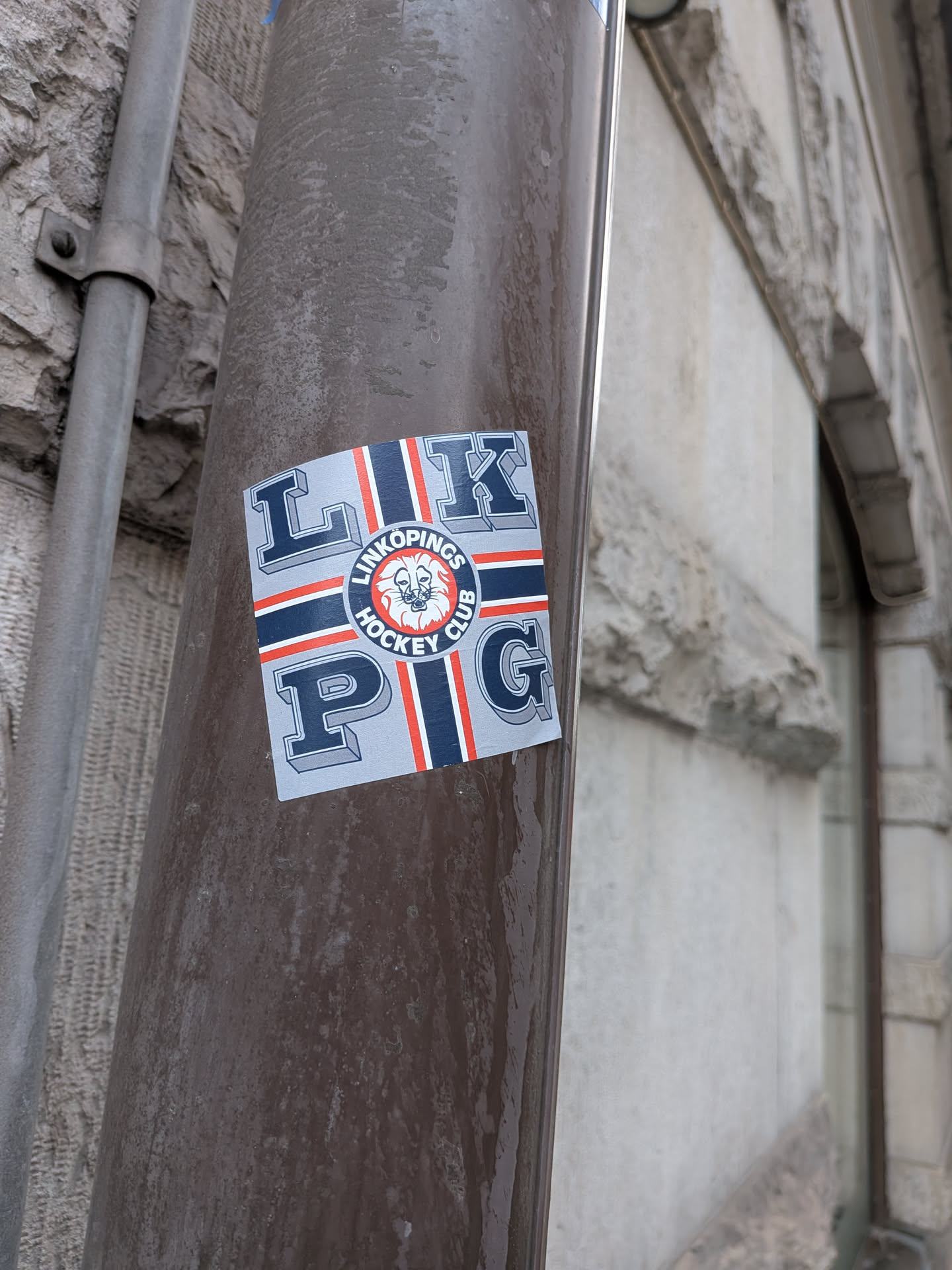 A square sticker placed on a brown painted metal pipe. It features the acronym 'LKPG' in stylized 3D serif block letters arranged across the corners on a light blue-grey field. Divided by a cross design composed of dark blue stripes bordered by white and red lines, the center bears the circular crest of Linköpings Hockey Club, displaying a roaring white lion head against a red center, encircled by a dark blue band reading 'LINKÖPINGS HOCKEY CLUB'.