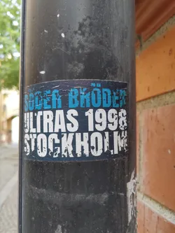 A worn sticker on a pole, featuring the text 'Söder Bröder Ultras 1998 Stockholm'. The sticker is predominantly black and white with blue accents. The text is slightly faded and damaged from wear.
