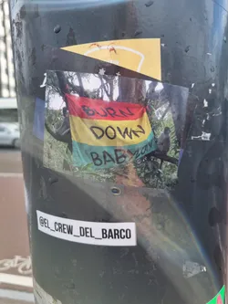 A photo of a sticker depicting two monkeys sitting on a tree branch with a flag that says "BURN DOWN BABYLON" in black and red text. The sticker is affixed to a pole. A smaller sticker underneath contains the text @el_crew_del_barco