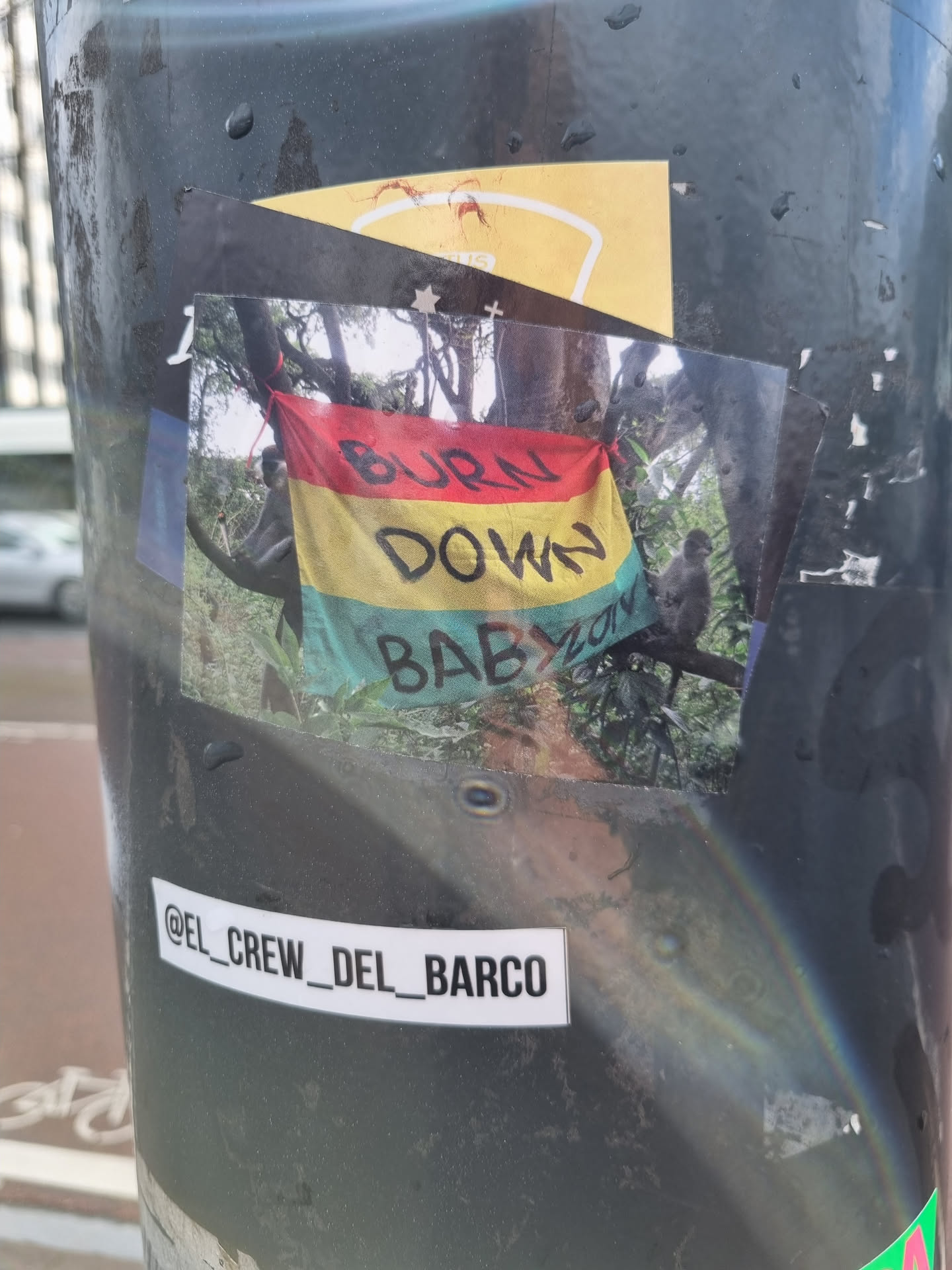 A photo of a sticker depicting two monkeys sitting on a tree branch with a flag that says "BURN DOWN BABYLON" in black and red text. The sticker is affixed to a pole. A smaller sticker underneath contains the text @el_crew_del_barco
