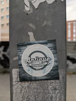 This square sticker is adhered to a gray pole. The design features a circle containing the text "Antique Barberia Peluqueria" in a stylish script, along with the location "Valencia". The background of the sticker has a textured wood-like appearance.
