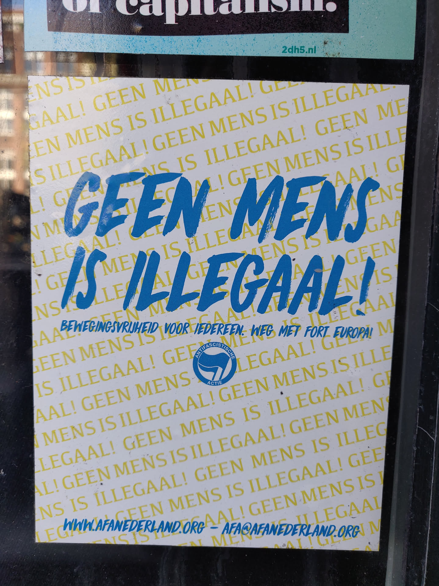 Sticker promoting the rights of migrants and refugees. It features the text "Geen mens is illegaal!" multiple times, along with the website www.afanederland.org and the logo of the organization BewegingSyruhieid voor iedereen. The sticker is meant to raise awareness of the issue and encourage people to support the cause.