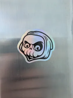 This is a holographic sticker of a skull. The skull has large, expressive eyes and a slightly menacing expression. The lines are thick and black, contrasting with the holographic background.