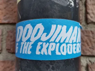 A light blue sticker with the text "Doojiman & The Exploders" written in a stylized white font. The sticker is applied to a dark-colored cylindrical object, possibly a pipe, against a brick wall background.