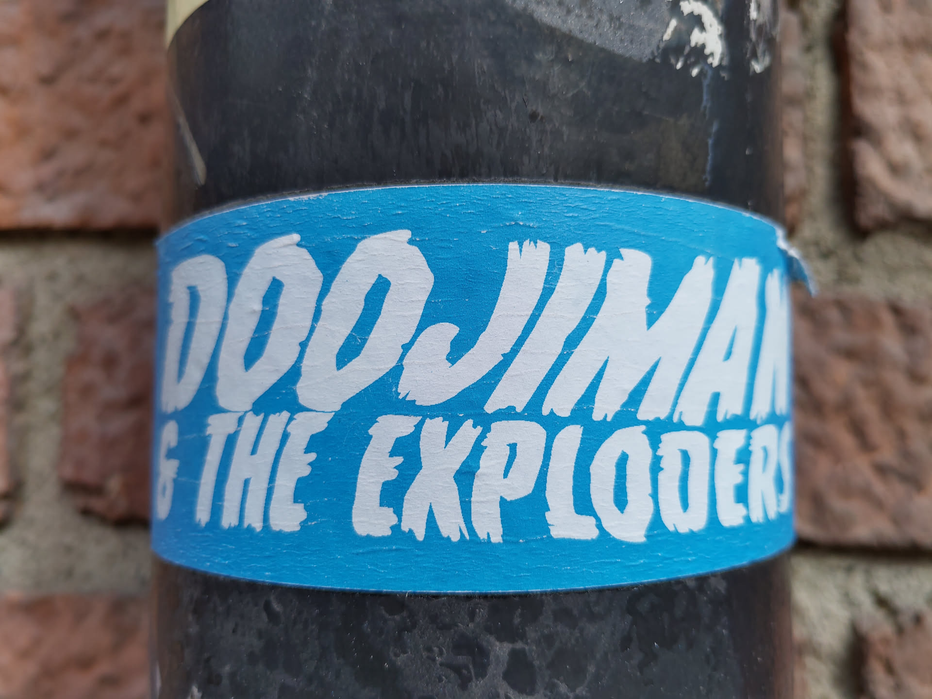 A light blue sticker with the text "Doojiman & The Exploders" written in a stylized white font. The sticker is applied to a dark-colored cylindrical object, possibly a pipe, against a brick wall background.