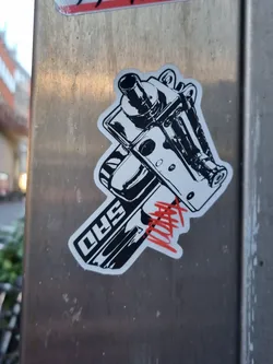 Black and white sticker depicting a detailed illustration of what looks like a car engine part or a similar mechanical component. The sticker has a stylized design with some red tagging on it and the letters "5HD" are visible on the bottom part of the illustration. The sticker is affixed to a metallic surface outdoors.
