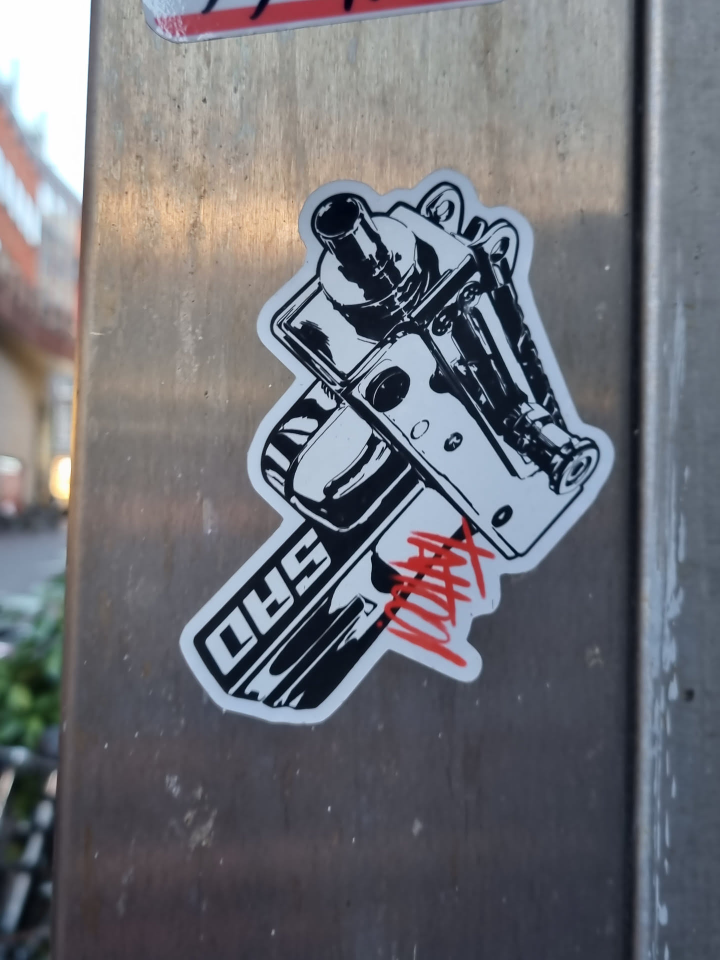 Black and white sticker depicting a detailed illustration of what looks like a car engine part or a similar mechanical component. The sticker has a stylized design with some red tagging on it and the letters "5HD" are visible on the bottom part of the illustration. The sticker is affixed to a metallic surface outdoors.